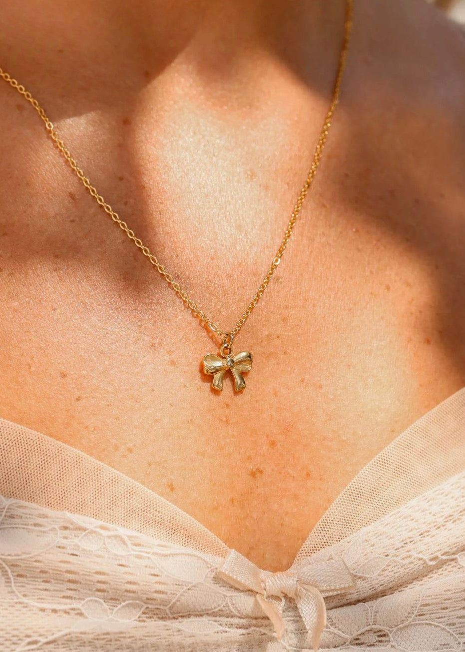 A close-up of a model's chest wearing a delicate gold chain necklace with a small bow-shaped pendant, paired with a cream lace camisole under warm natural sunlight.